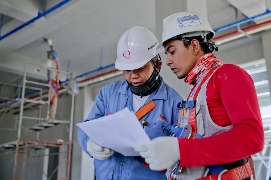 Two construction workers in safety gear reviewing documents inside a building under construction.