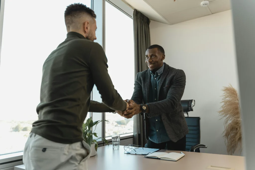 Two men in business casual attire shaking hands in a bright, modern office with a large window in the background.