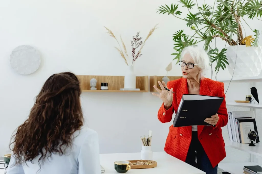 A woman with white hair in a bright red blazer holds a black binder while gesturing during a conversation in a modern, plant-filled office.