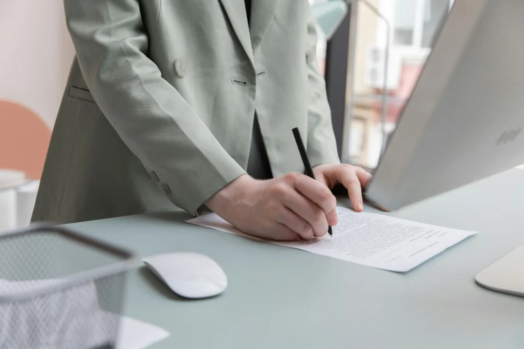 Person in green blazer signing document at desk with computer and office supplies