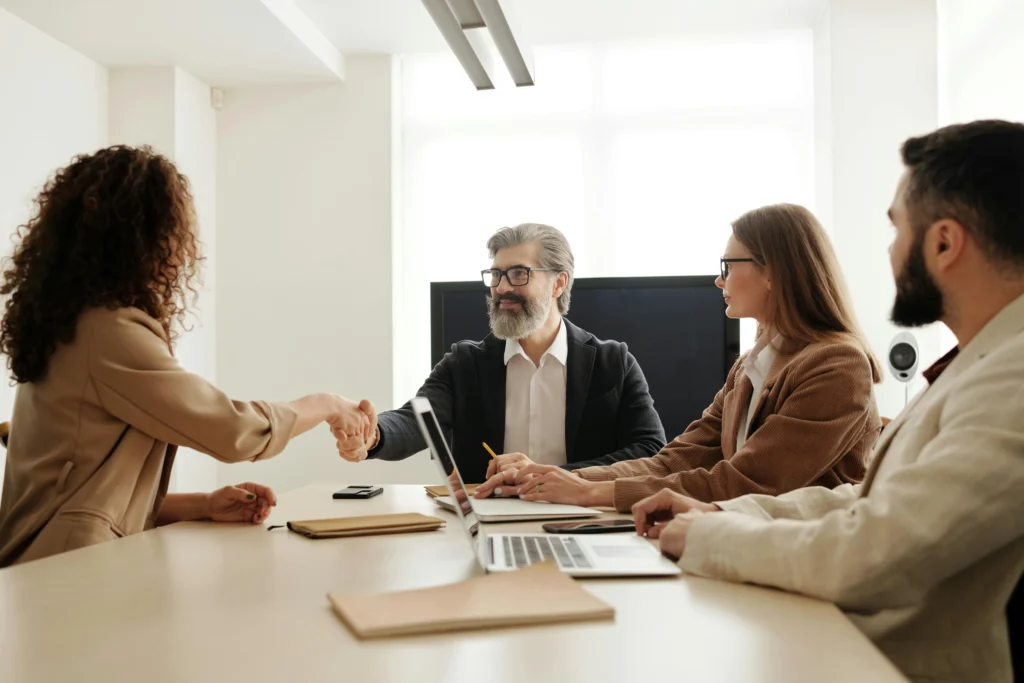 A man with a grey beard looks on as two colleagues shake hands across a conference table during a meeting.
