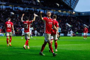 Football players in red kits celebrate on a stadium field during a match, with a cheering crowd in the stands in the background.
