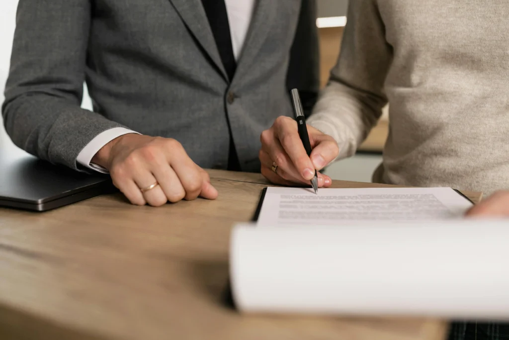Close-up of two people at a wooden desk signing a paper document with a black pen.