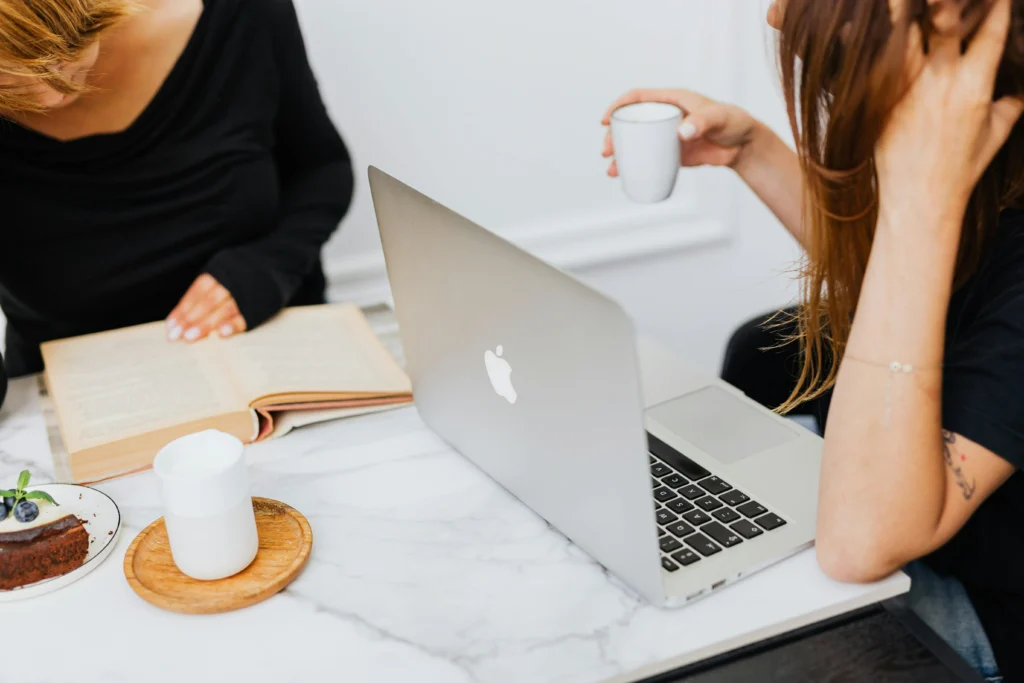 High-angle view of two women at a marble table with a laptop, an open book, coffee, and a dessert.