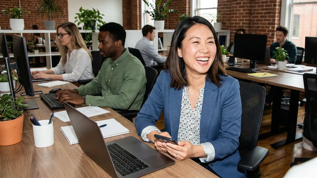 A smiling professional holding a smartphone while sitting at a laptop with colleagues in a modern office, illustrating the engaged workforce supported by the right HR software for growing companies.