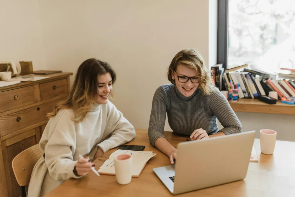 Two women smiling and looking at a laptop screen while sitting at a wooden table with notebooks and coffee.