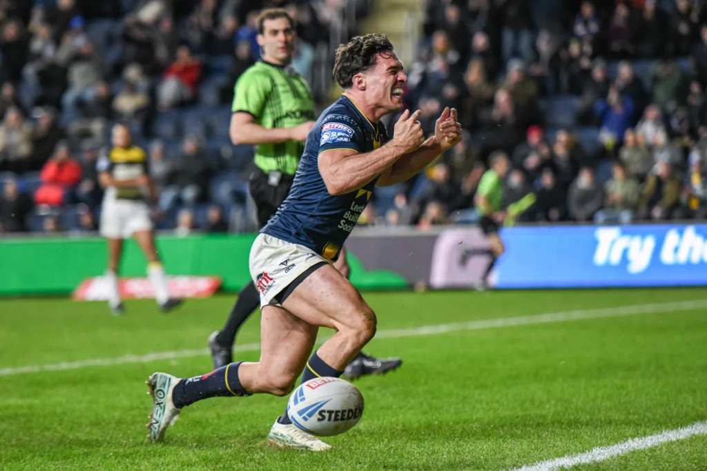 A rugby player in a navy uniform exuberantly clutches the ball, celebrating a try. The background shows a blurred referee, players, and a cheering crowd.