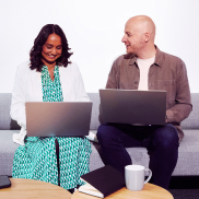 Two people sitting on a sofa, working on laptops. They are smiling and appear collaborative. A notebook and coffee mug are on the table in front of them.