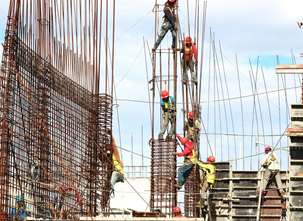 Construction workers wearing safety gear climb and work on rebar structures against a clear sky, conveying teamwork and progress on a building site.