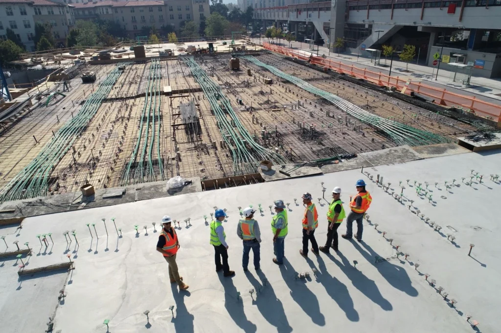 Seven construction workers wearing safety vests and helmets stand on a large building site covered in metal rods and pipes under sunny skies.