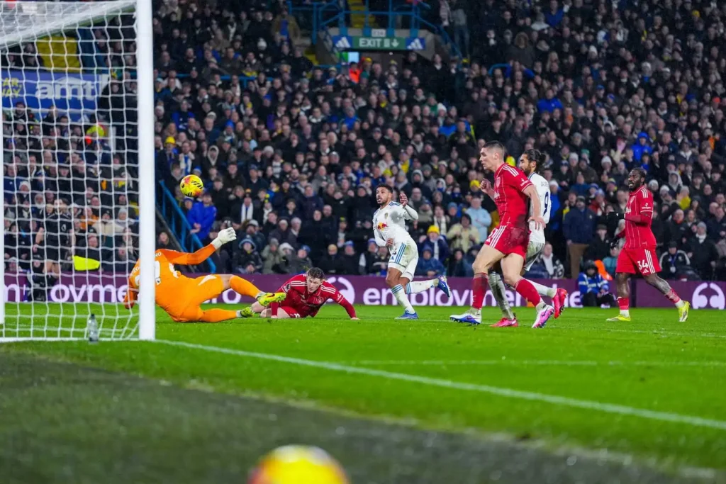 A soccer match moment with a goalkeeper in orange diving to save a ball nearing the goal. Players in red and white, with a cheering crowd, create tense action.