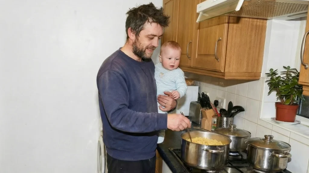 A smiling father holds his baby in one arm while stirring a pot on the stove, representing working parents and the importance of updated UK family leave policies and shared parental leave coming in April 2026.