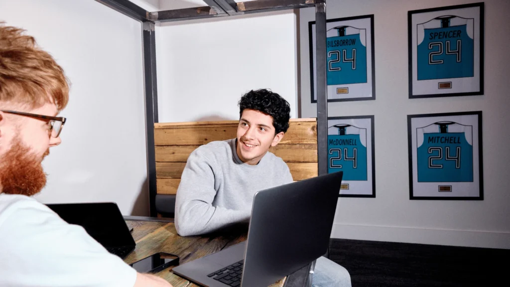 Two people sit at a table with laptops, smiling and engaged in conversation. Behind them, framed sports jerseys with the number 24 are displayed on the wall.