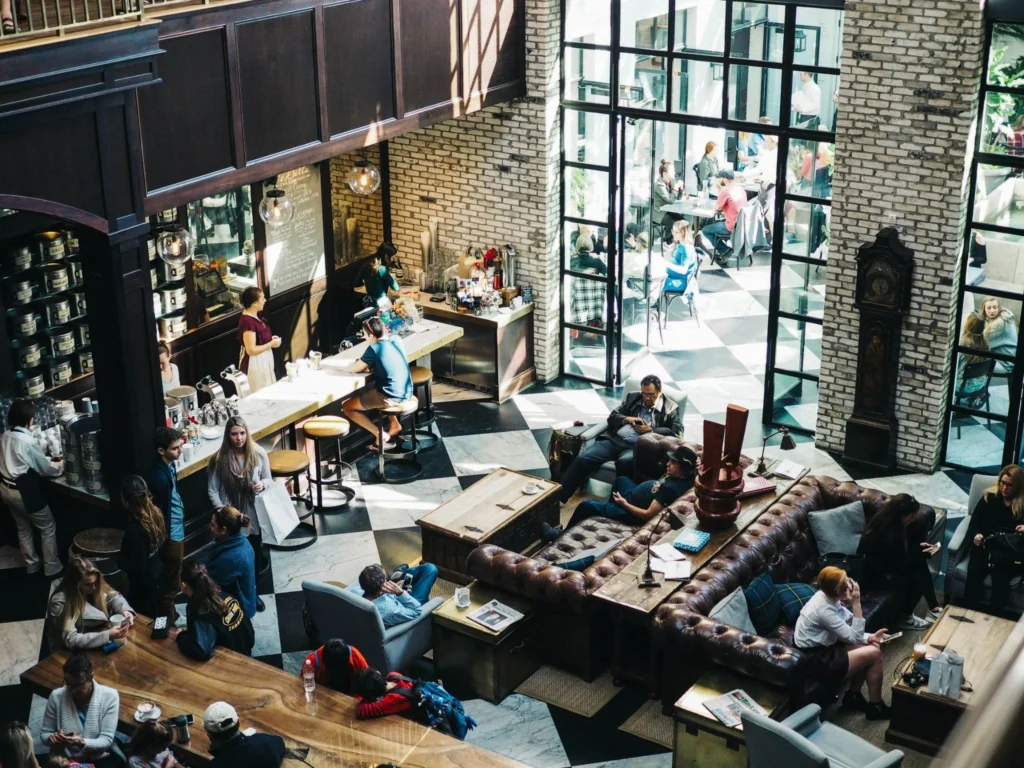 A bustling café with people chatting and relaxing, featuring cozy leather sofas, a long counter with barstools, and large windows letting in sunlight.