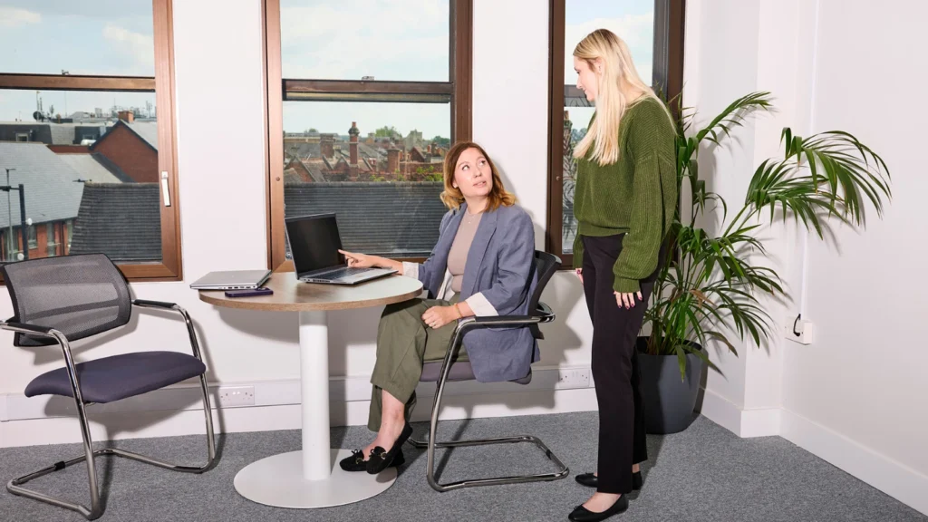Two women in an office beside large windows; one seated at a table with a laptop, the other standing, engaged in conversation. A potted plant adds greenery.