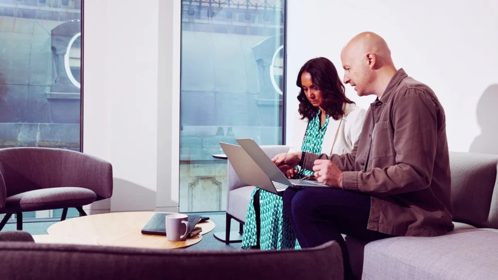 A man and woman sit on a sofa in a modern office, discussing work on a laptop. The atmosphere is collaborative and focused. A window and table with a mug are in the background.