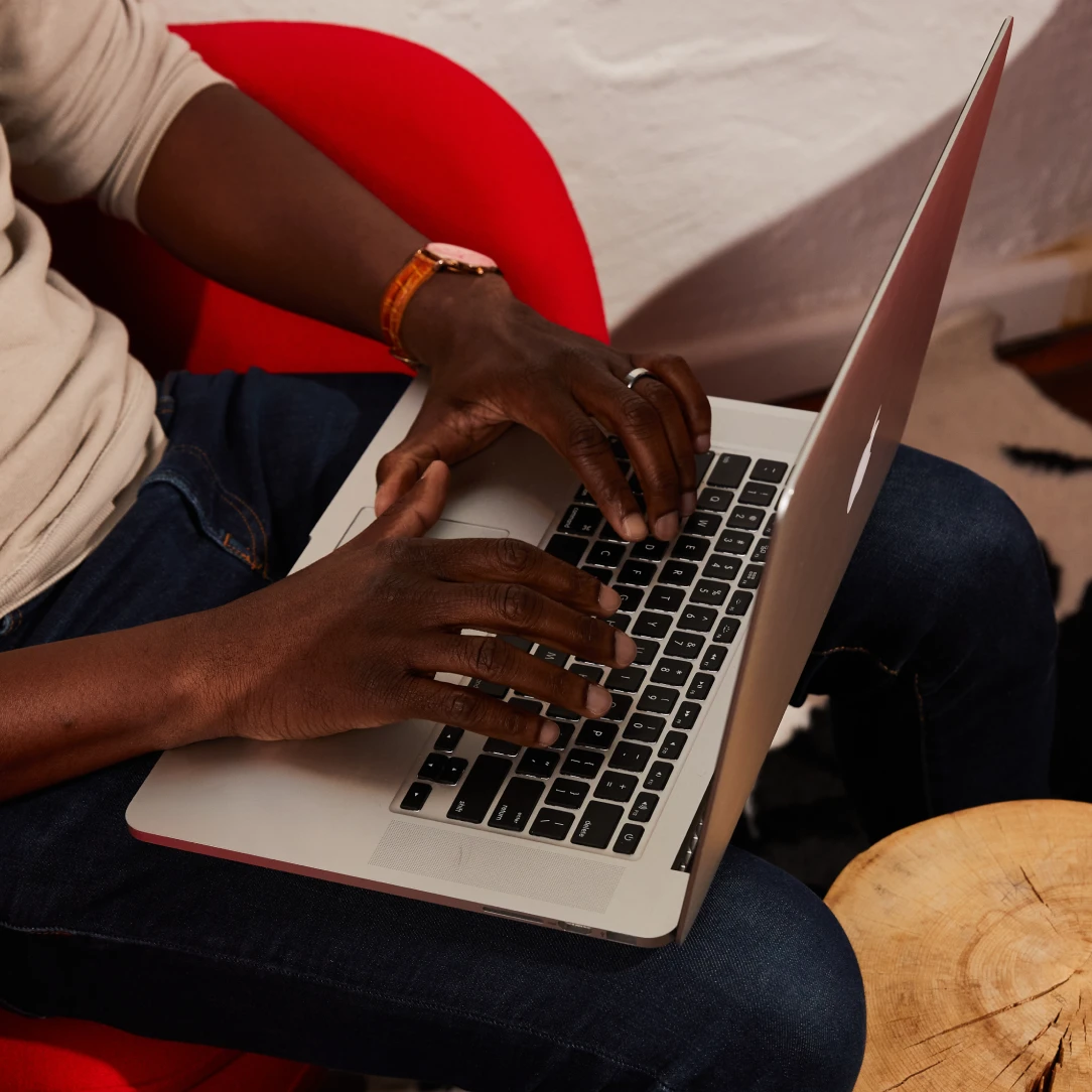 A person in a beige sweater and jeans types on a laptop while seated in a red chair. 
