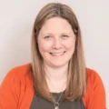 Smiling woman with long, light brown hair, wearing an orange cardigan over a dark shirt and a necklace, against a neutral background. Warm and friendly tone.