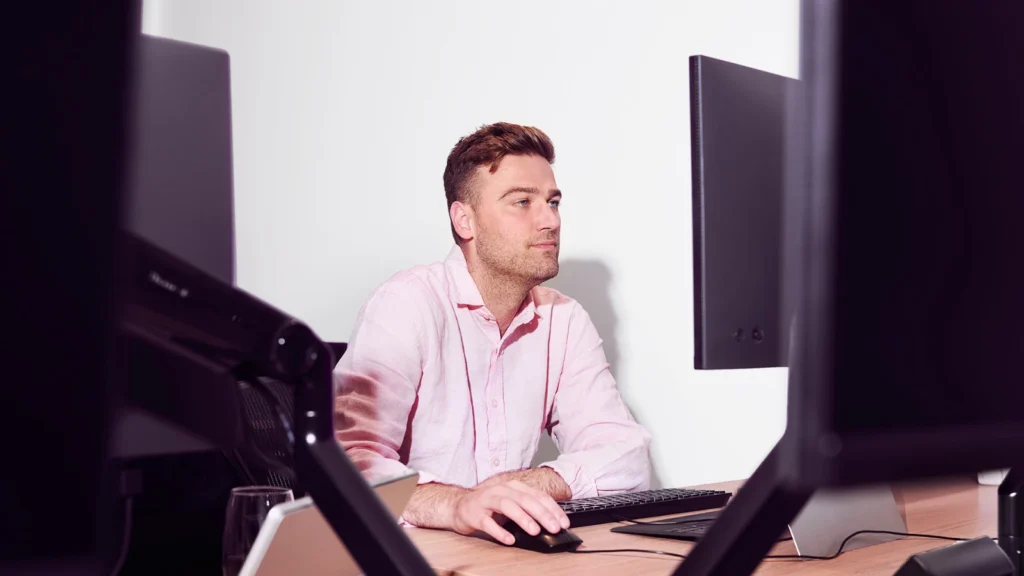 A man in a pink shirt sits focused at a desk with multiple monitors, exuding concentration and professionalism in a modern office setting.