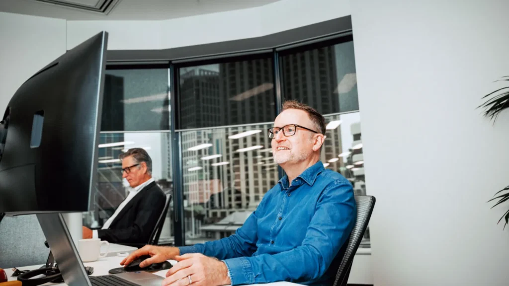 A smiling male professional wearing glasses and a blue shirt working at his computer in a modern corporate office, illustrating how UK employers can streamline their operations using business process automation.