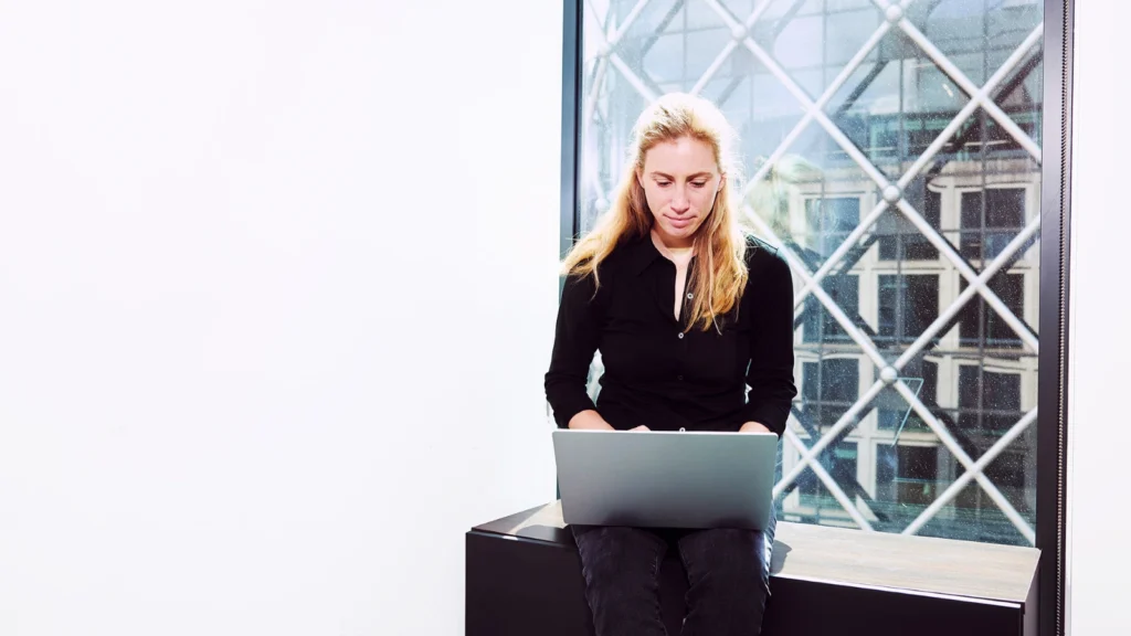 A woman in a black shirt sits on a ledge by a large window, working on a laptop. The window shows a crisscross pattern with a building view outside.