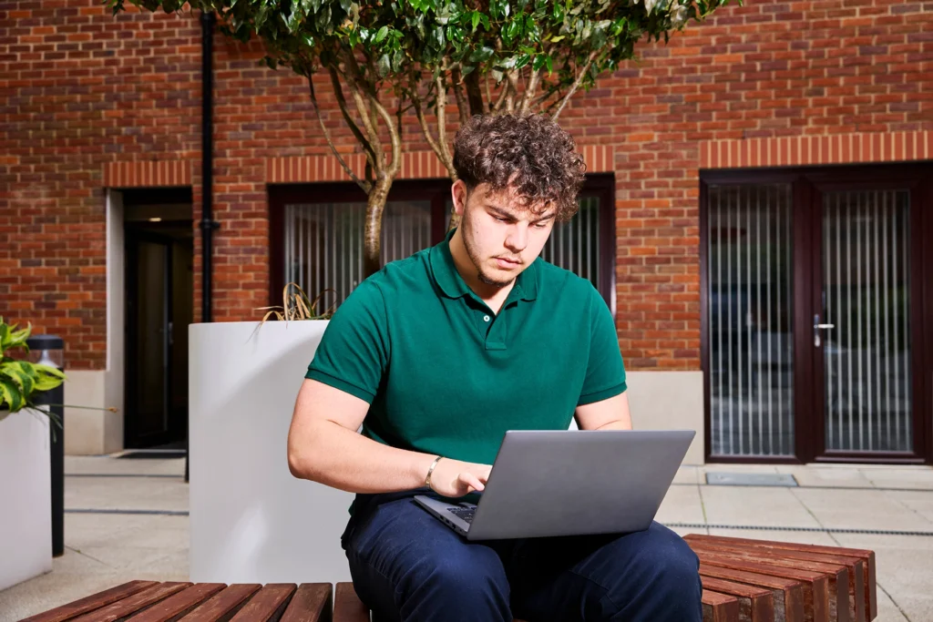 A man seated on a bench, focused on his laptop, surrounded by greenery in a park setting.