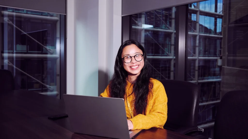 Smiling person wearing a yellow sweater and glasses sits at a laptop in a modern office with large windows, conveying a professional and cheerful mood.