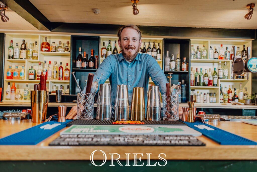 Man in blue shirt standing behind a bar lined with bottles of spirits and cocktail equipment