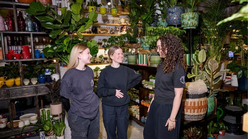 Three people in a plant store, surrounded by lush green plants and colorful pots. They are smiling and engaged in friendly conversation, conveying a warm, cheerful atmosphere.