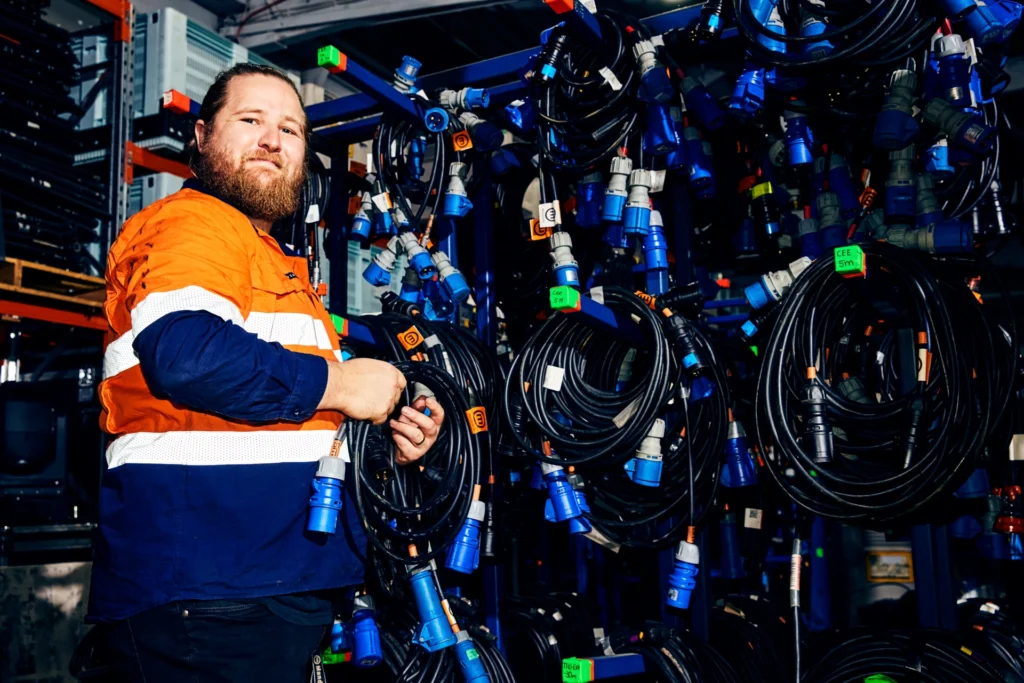A man wearing a hi-vis orange shirt standing in a workshop surrounded by organized racks of black and blue power cables.