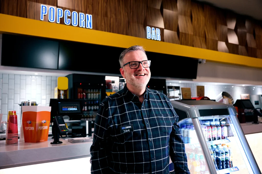 A smiling man named Dave wearing a checkered shirt and glasses standing in front of a cinema concession popcorn bar.