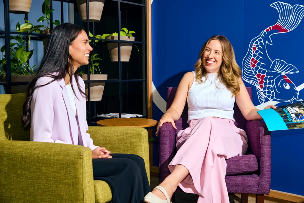 Two women smiling and chatting while seated in armchair seats in a brightly decorated office lounge area.