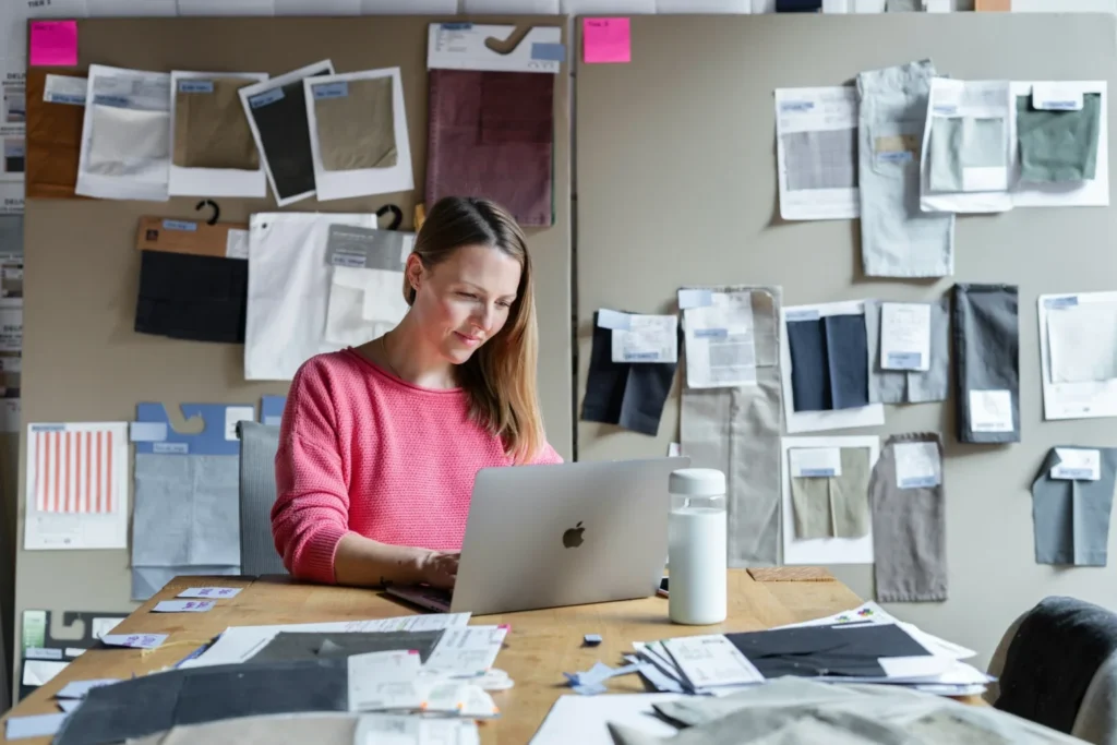 A designer wearing a pink sweater sits at a workspace typing on a laptop in front of fabric swatch boards.