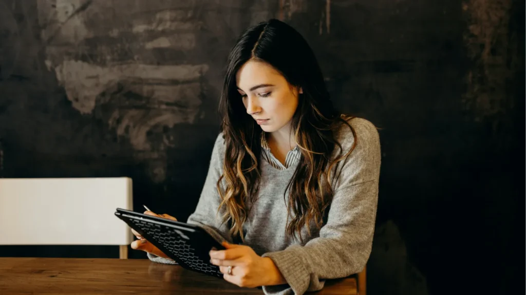 A woman with long wavy hair wearing a grey sweater sits at a wooden table, focused on using a stylus to write or draw on a tablet device.