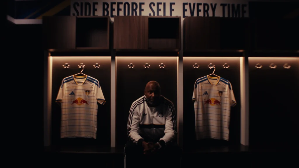 Lucas Radebe sits in a stadium locker room flanked by hanging Leeds United jerseys under "SIDE BEFORE SELF EVERY TIME".