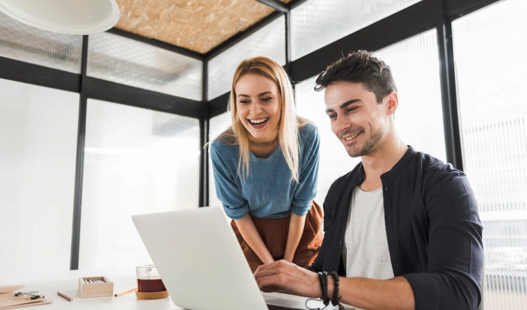 A smiling woman stands leaning over the shoulder of a male colleague sitting at a desk as they look together at a laptop screen.