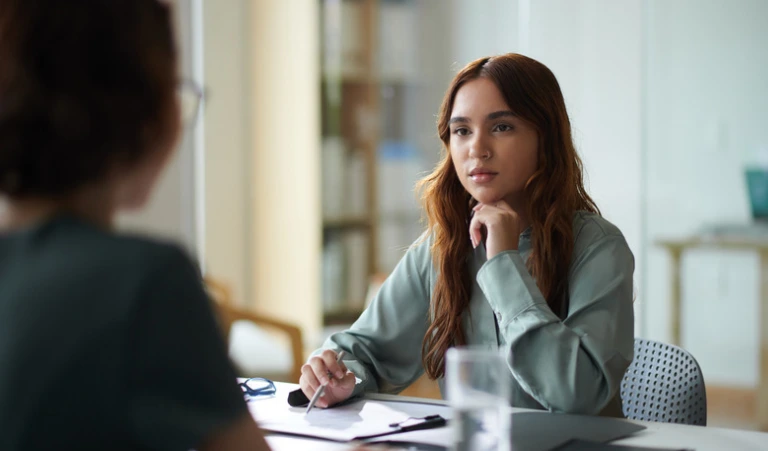 An attentive employee rests her hand on her chin as she looks across a desk at her manager during a formal meeting or performance review