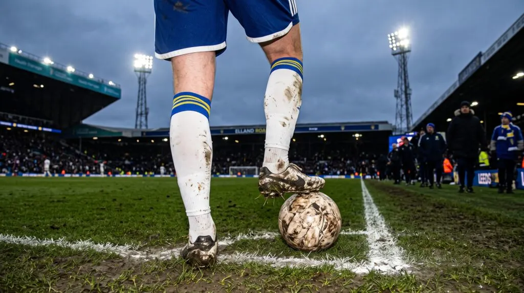 A soccer player in mud-splattered socks stands at the corner of a stadium field with one foot resting on a muddy ball.