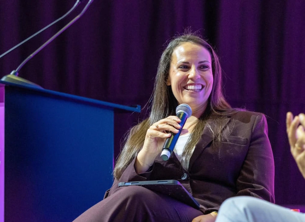 A woman in a dark blazer smiles while speaking into a microphone during a panel event stage discussion.
