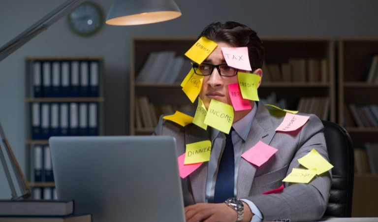 An overwhelmed office worker sits at a laptop late at night with colorful sticky notes covering his face, glasses, and suit jacket.