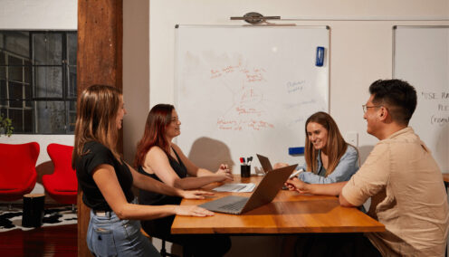 Group of colleagues around a desk chatting in their office in front of a whiteboard.