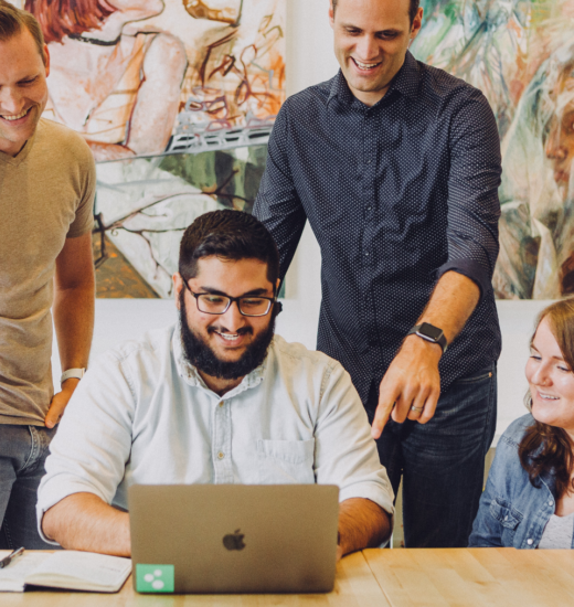 Man sits at a table on his laptop, while his colleagues surround him observing his work