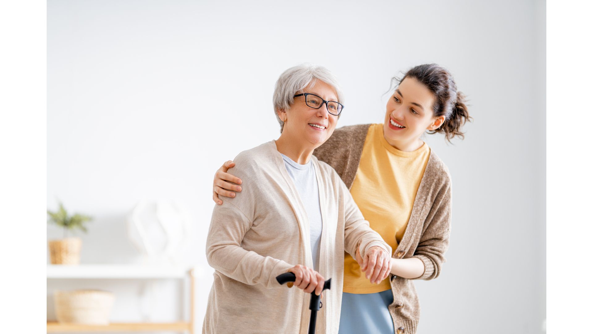 A younger woman smiles as she supports an older woman with grey hair who is using a walking cane