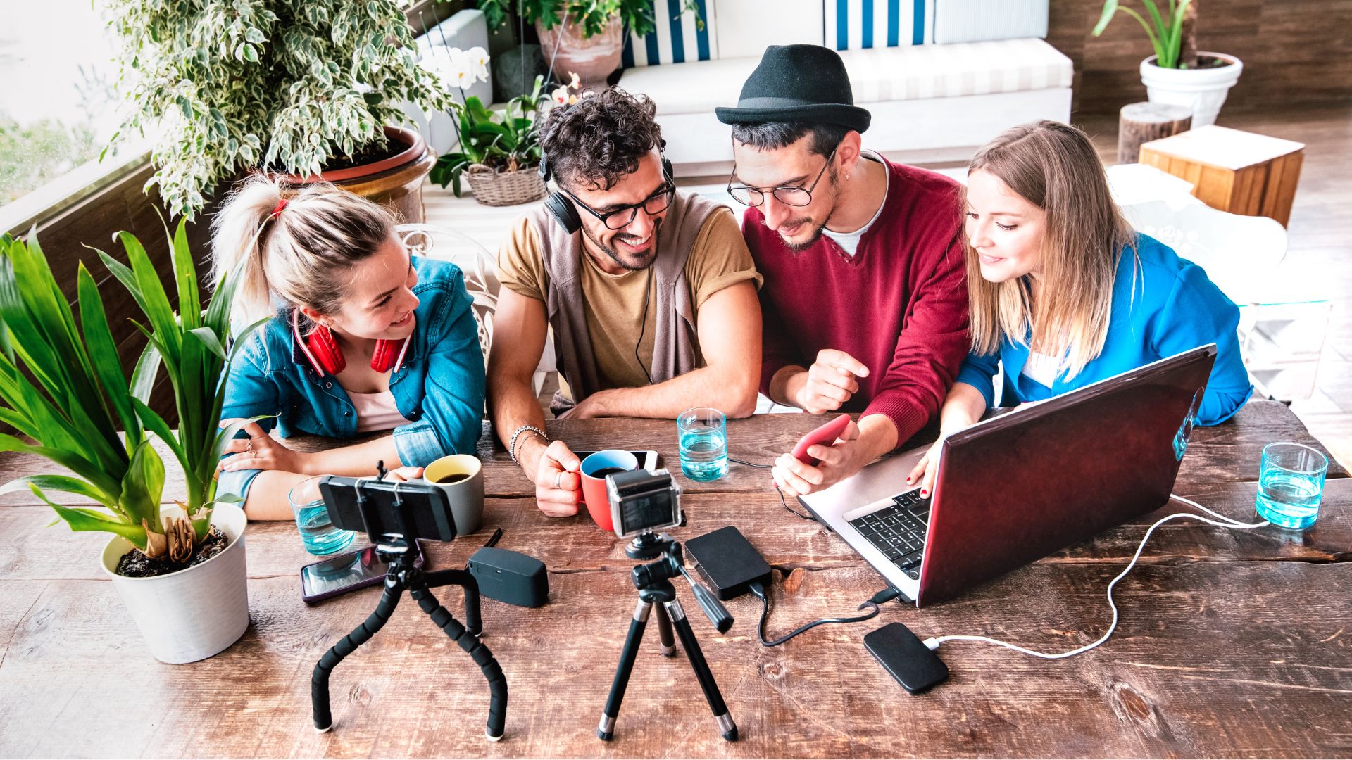 A group of four young adults gather around a table, smiling and working together with a laptop, smartphone, and camera equipment