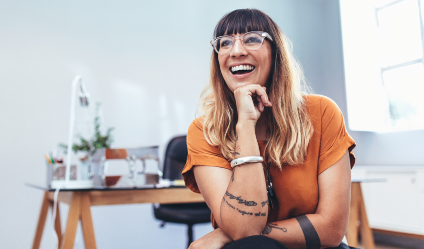 Women wearing glasses and sitting at her desk with one hand under her chin and a friendly smile.