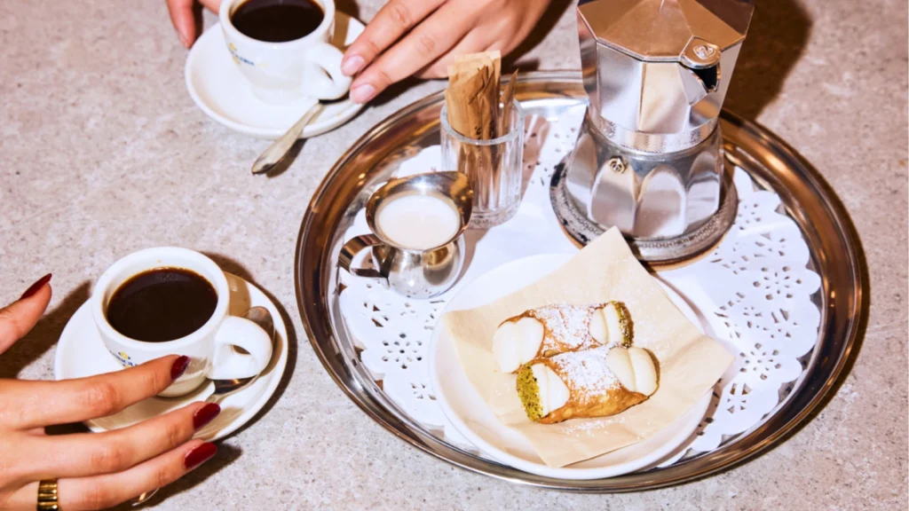 A top-down view of an espresso spread on a speckled grey table. Two pairs of hands are shown touching small white cups of black coffee on saucers. In the center, a round silver tray holds a classic silver moka pot, a small metal creamer pitcher, a glass holding sugar packets, and two small cannoli dusted with powdered sugar on a napkin-lined white plate.