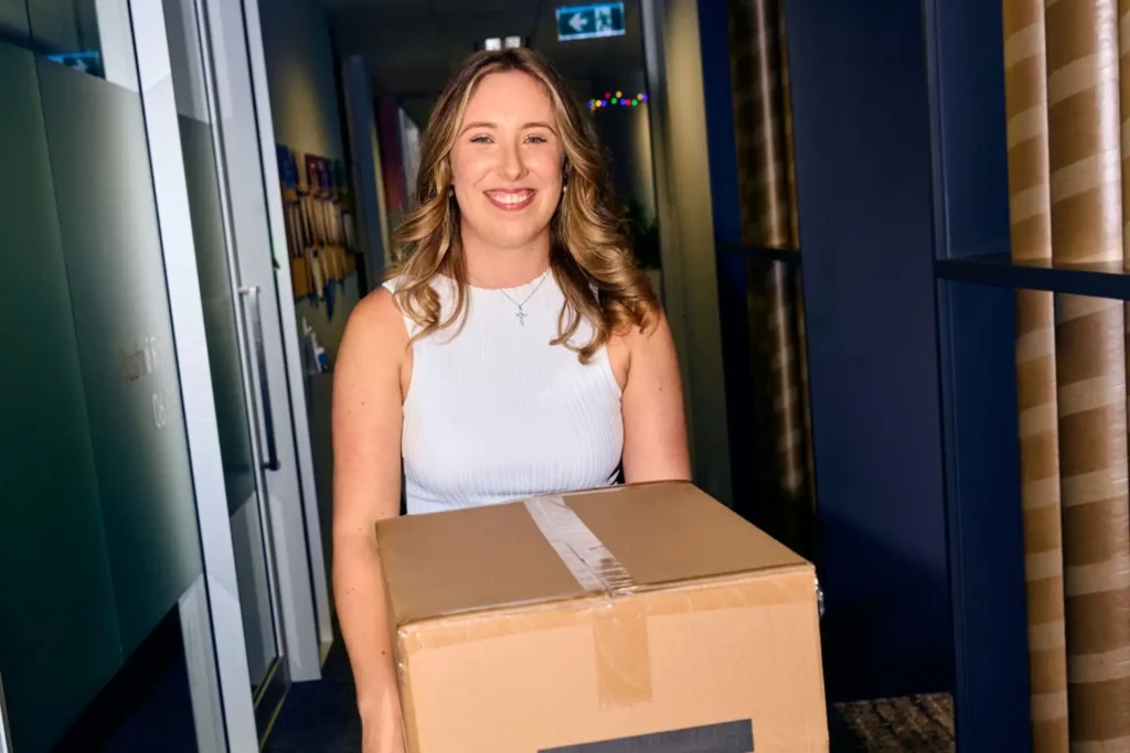 Smiling woman in a white sleeveless top carries a large cardboard box in a brightly lit hallway. The setting appears to be an office, conveying a cheerful mood.