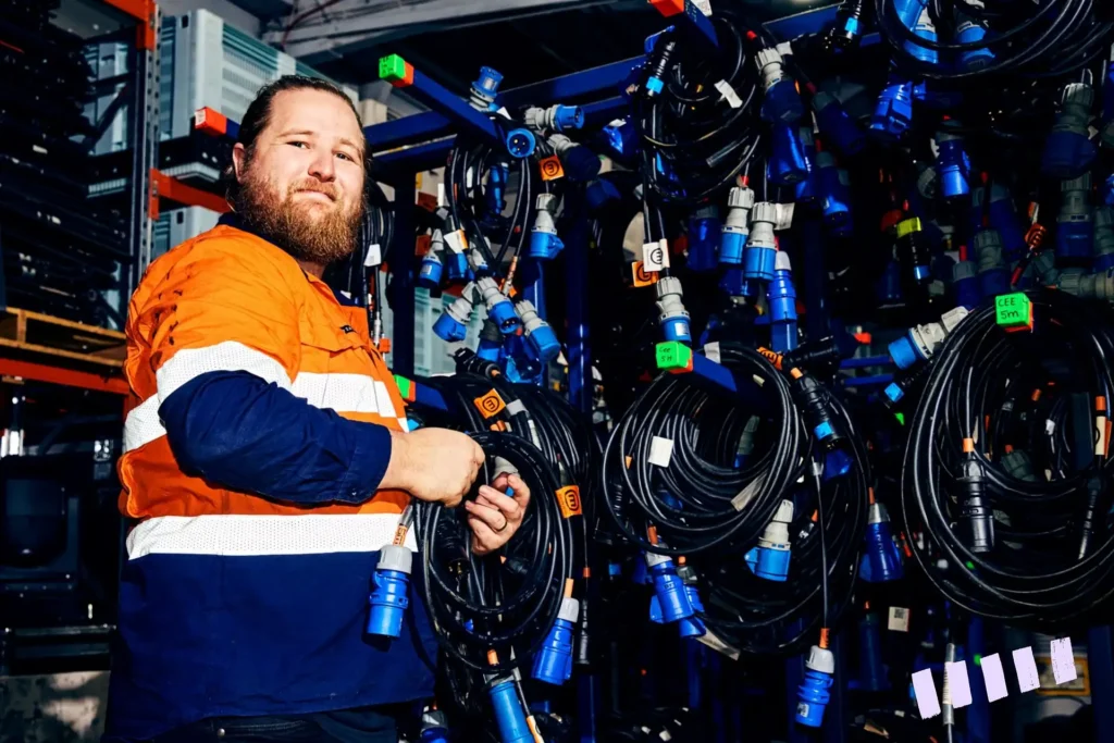 A man in an orange and navy uniform handles coiled cables in a warehouse filled with neatly hung cables. The setting is organized and industrious.