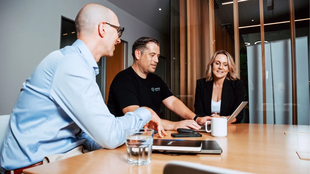 Three colleagues seated around a wooden conference table discussing information on a open laptop screen.