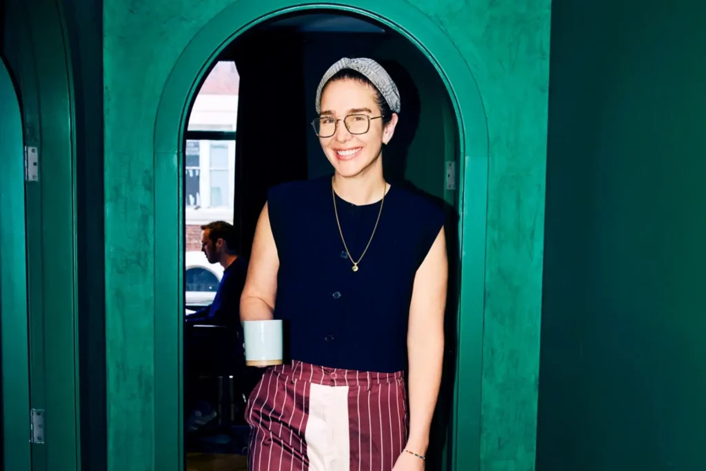 Person smiling in a stylish cafe, wearing glasses, a sleeveless top, striped pants, and a headband, holding a mug. Green arched doorway in the background.